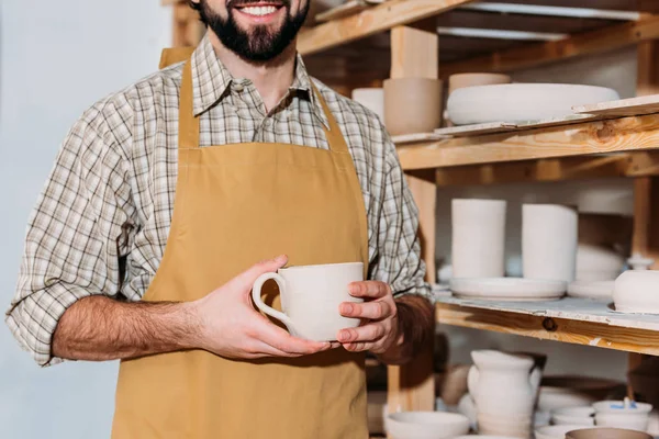 Cropped view of male potter holding ceramic cup and standing near shelves with dishware — Stock Photo
