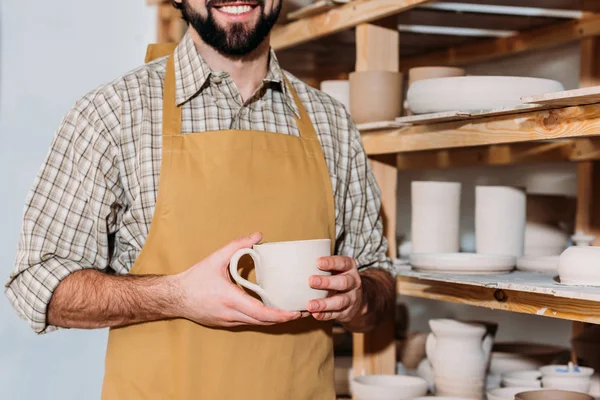 Vista ritagliata di vasaio maschile in possesso di tazza di ceramica e in piedi vicino a scaffali con stoviglie — Foto stock