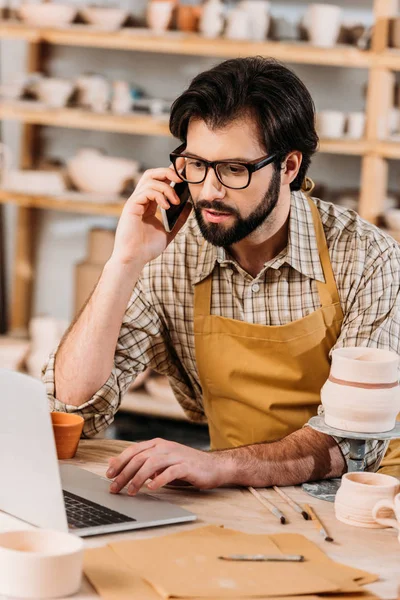 Hombre hablando en el teléfono inteligente mientras trabaja en el ordenador portátil en taller de cerámica - foto de stock