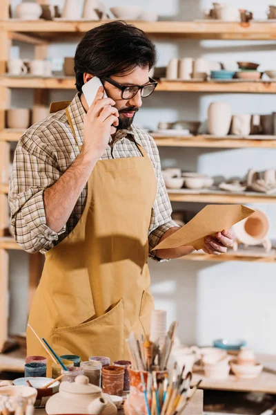 Alfarero hablando en el teléfono inteligente mientras busca en el sobre en el taller — Stock Photo