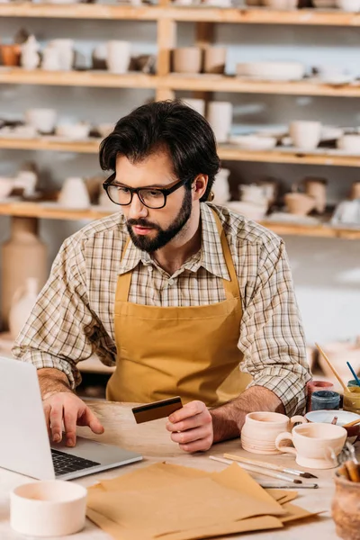 Bearded man shopping online with laptop and credit card in pottery workshop — Stock Photo