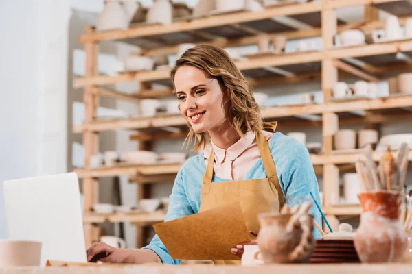 Femme souriante blonde avec ordinateur portable et document en atelier de poterie — Photo de stock