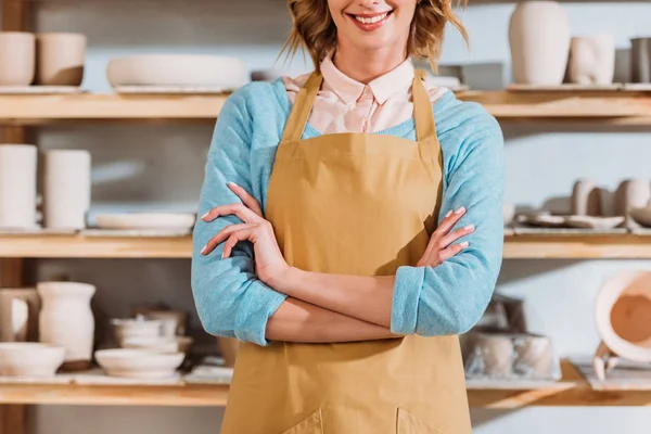 Cropped view of potter with crossed arms standing near shelves with ceramic dishware — Stock Photo