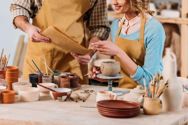 Cropped view of potters with papers, ceramics, paints and brushes in workshop — Stock Photo