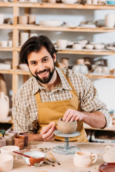 Ceramista sorridente in grembiule guardando la macchina fotografica e decorando ceramiche in officina — Foto stock