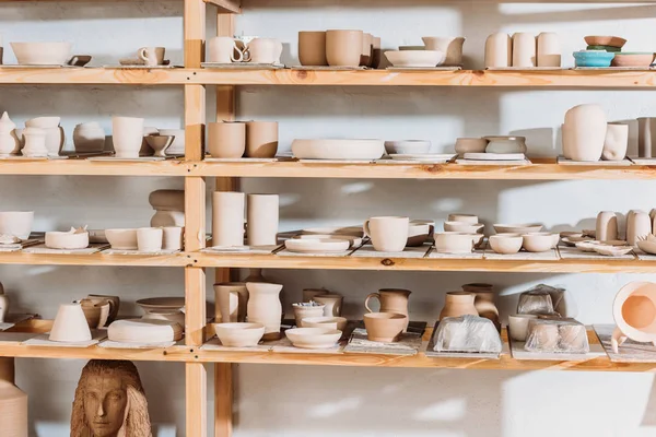 Different ceramic dishware on wooden shelves in pottery workshop — Stock Photo