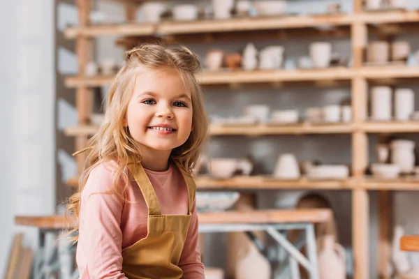Bionda sorridente bambino guardando la fotocamera in laboratorio di ceramica — Foto stock