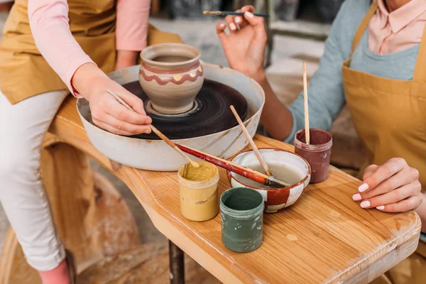 Cropped view of teacher and kid painting ceramic pot with brushes in workshop — Stock Photo