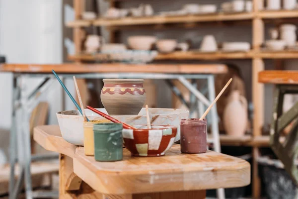 Ceramic pot, brushes and paints on wooden table in pottery workshop — Stock Photo