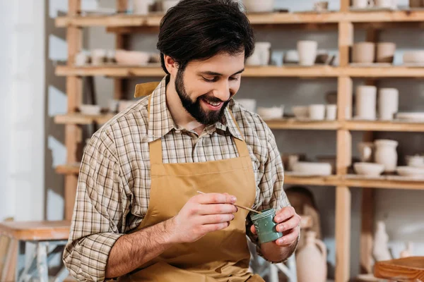 Sonriente alfarero macho sosteniendo pintura y pincel en taller - foto de stock