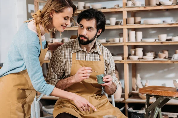 Alfarero mujer enseñando barbudo hombre cómo pintar cerámica en taller - foto de stock