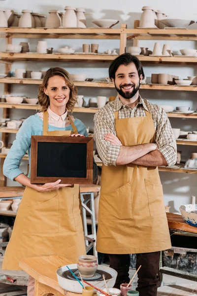 Two owners holding chalkboard in pottery workshop with ceramics on shelves — Stock Photo