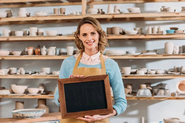 Beau propriétaire souriant avec tableau noir en atelier de poterie — Photo de stock