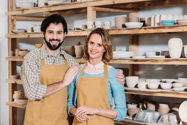 Couple in aprons hugging in pottery workshop with ceramics on shelves — Stock Photo