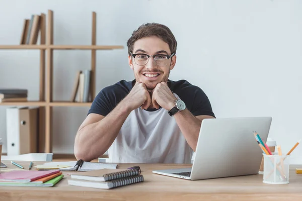 Estudiante alegre sentado en la mesa con cuadernos y portátil - foto de stock