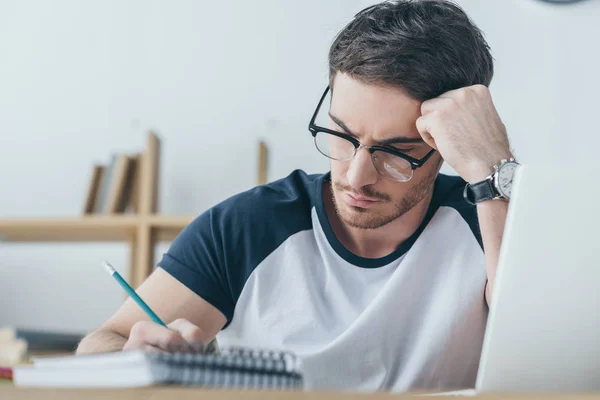 Schöner männlicher Student mit Brille, der in ein Werbebuch schreibt — Stockfoto