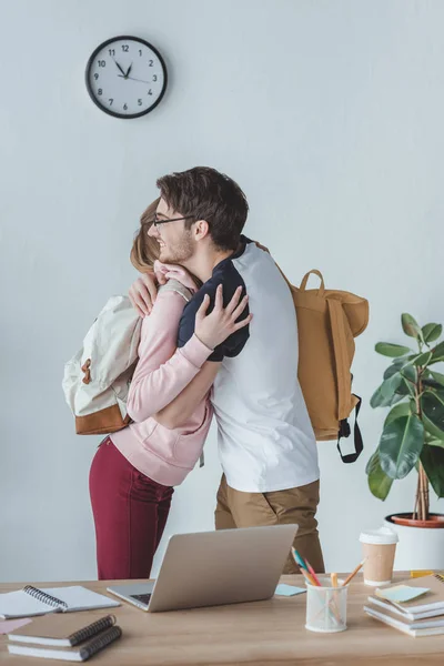 Friends with backpacks hugging, books and laptop standing on table — Stock Photo