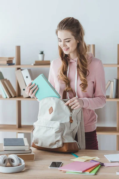 Привлекательная улыбающаяся девушка кладет книгу в рюкзак — стоковое фото
