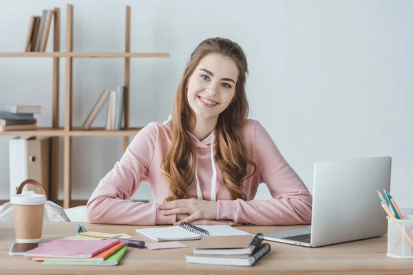 Estudiante sonriente sentada en la mesa con portátil y copybooks - foto de stock