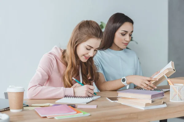 Niñas multiétnicas que estudian juntos y escriben en copybook y libro de lectura en la mesa - foto de stock