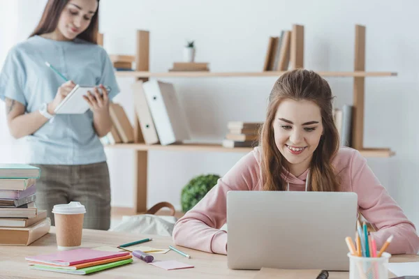 Chica rubia estudiando con el ordenador portátil, mientras que su amigo escribiendo en copybook detrás - foto de stock