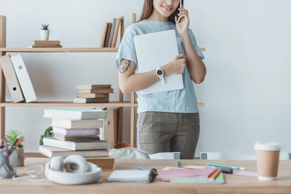 Cropped view of student holding laptop while talking on smartphone — Stock Photo