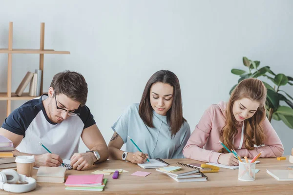 Giovani studenti multiculturali che scrivono in quaderni a tavola — Foto stock