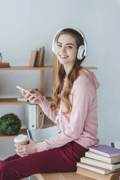 Estudiante rubia sonriente con café escuchando música con smartphone y auriculares - foto de stock