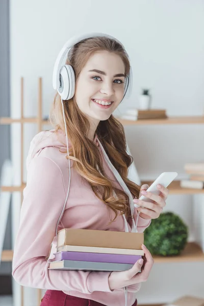 Étudiante souriante tenant des livres et écoutant de la musique avec smartphone et écouteurs — Photo de stock