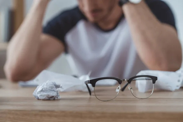 Selective focus of depressed student at table with crumpled papers and eyeglasses on foreground — Stock Photo