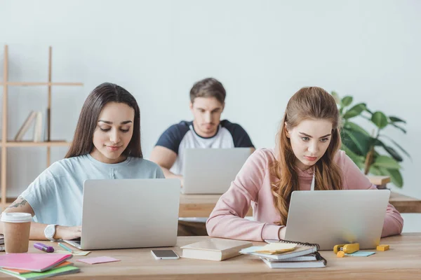 Jóvenes estudiantes multiétnicos sentados en las mesas y estudiando con ordenadores portátiles - foto de stock