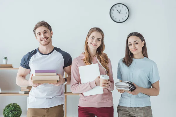 Estudiantes sonrientes sosteniendo libros, laptop, café y copybooks - foto de stock