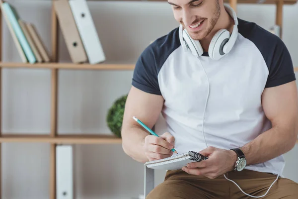 Smiling male student with headphones writing in notebook — Stock Photo