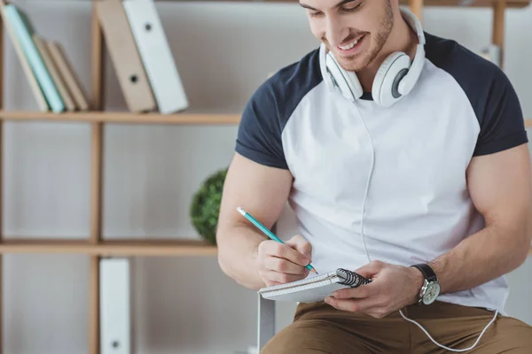 Étudiant souriant de sexe masculin avec écouteurs écrit dans un cahier — Photo de stock