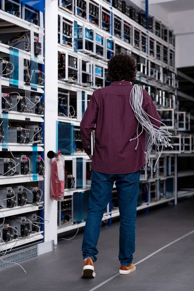 Vista trasera del ingeniero informático con cables de Ethernet en el hombro en la granja de minería criptomoneda - foto de stock