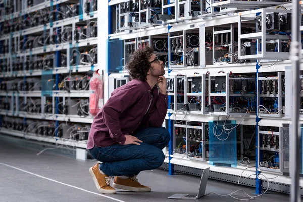 Молодой компьютерный инженер сидит на полу и смотрит на криптовалютную горнодобывающую ферму — стоковое фото