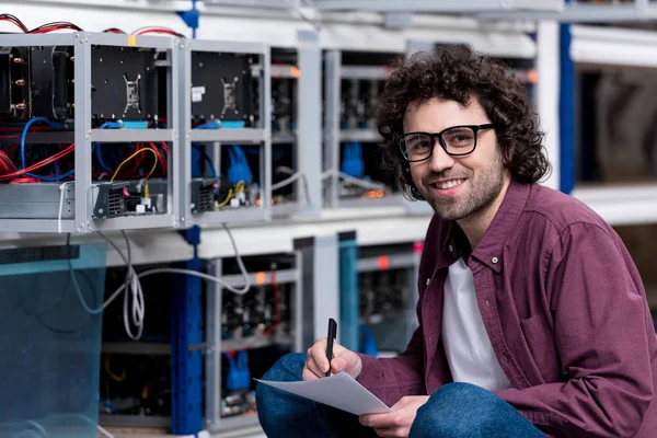 Joven ingeniero informático sonriente sentado en el suelo y escribiendo en la granja minera criptomoneda - foto de stock