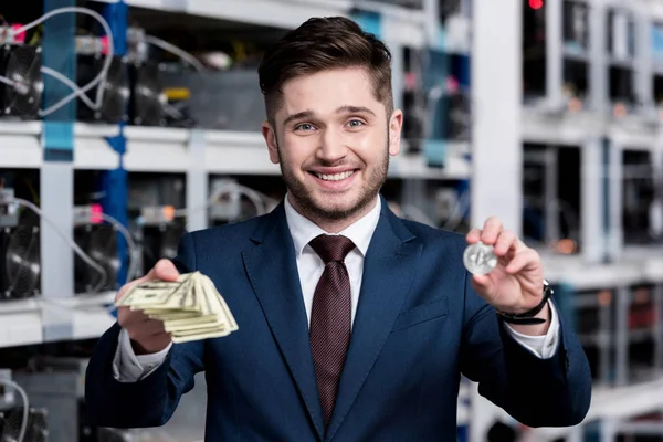 Apuesto joven empresario sosteniendo pila de dinero en efectivo y bitcoin en la granja minera criptomoneda - foto de stock
