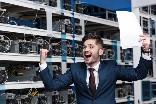 Glücklicher junger Geschäftsmann mit Papieren feiert Sieg auf Kryptowährungsfarm — Stockfoto