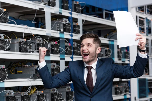 Feliz joven empresario con papeles celebrando la victoria en la granja minera criptomoneda - foto de stock