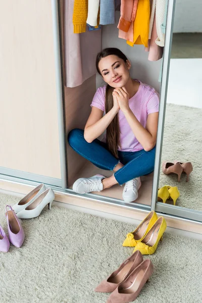 Attractive young woman sitting inside of cabinet with various shoes on floor at home — Stock Photo