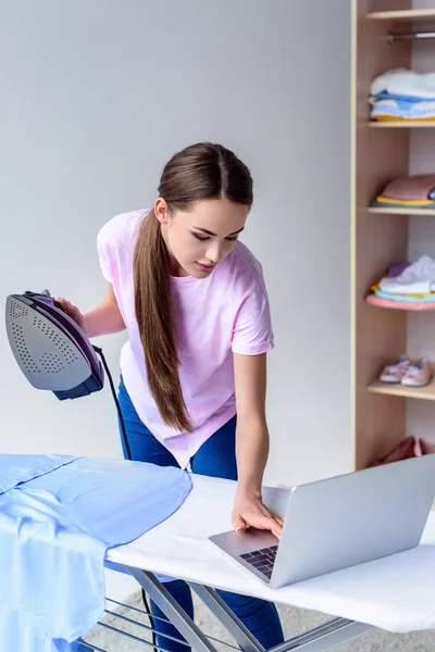 Young woman ironing clothing and working with laptop at home — Stock Photo