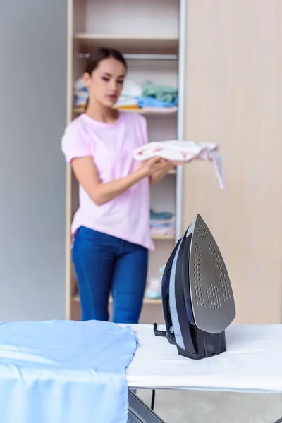 Bügeleisen an Bord mit verschwommener Frau im Hintergrund, die gefaltete Kleidung zu Hause hält — Stockfoto