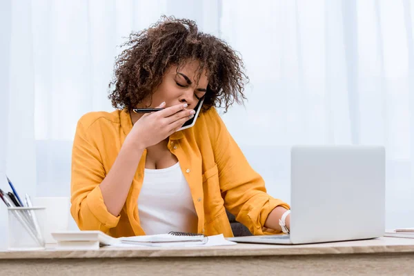 Exhausted young woman talking by phone at workplace and yawning — Stock Photo