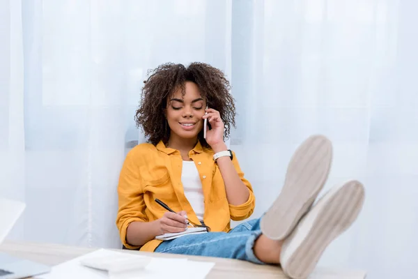 Happy mixed race woman talking by phone and writing in notebook — Stock Photo
