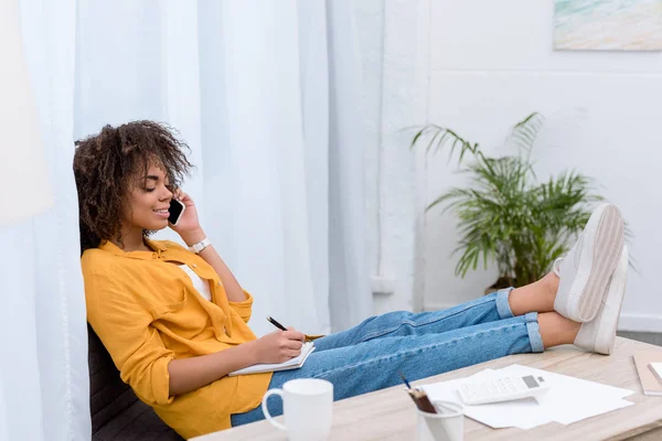 Belle jeune femme parlant par téléphone et écrivant dans un cahier — Photo de stock