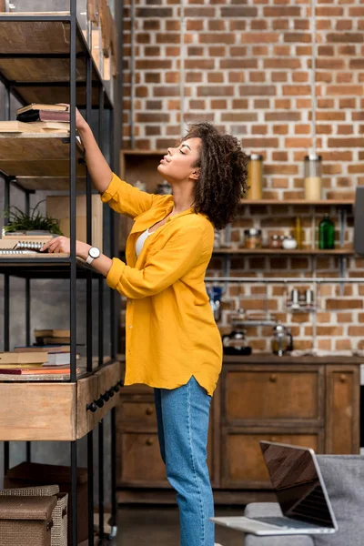 Beautiful young woman taking book from shelf at home — Stock Photo