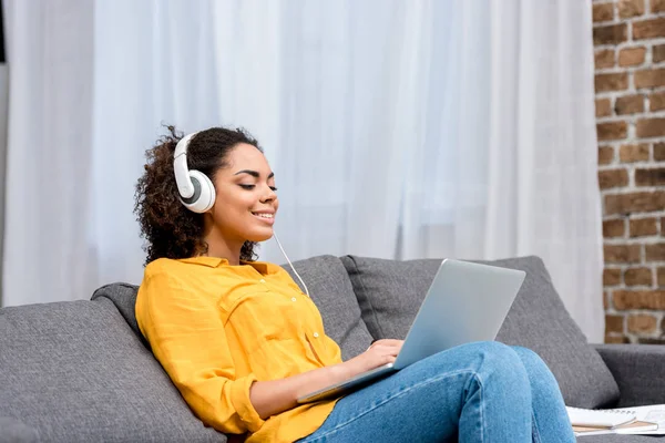 Sorrindo mulher afro-americana trabalhando com laptop e ouvindo música no sofá em casa — Fotografia de Stock