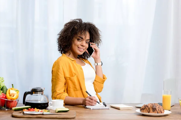 Souriant jeune femme parlant par téléphone à la cuisine et regardant la caméra — Photo de stock