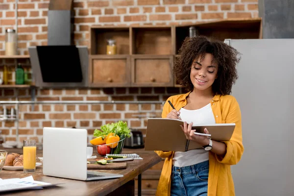 Bela jovem mulher trabalhando na cozinha e escrevendo em notebook — Fotografia de Stock